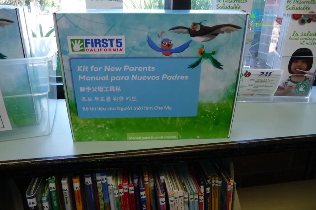 A First 5 California "Kit for New Parents" on a library shelf, labeled in English, Spanish, Chinese, Korean, and Vietnamese, above a row of children's books.