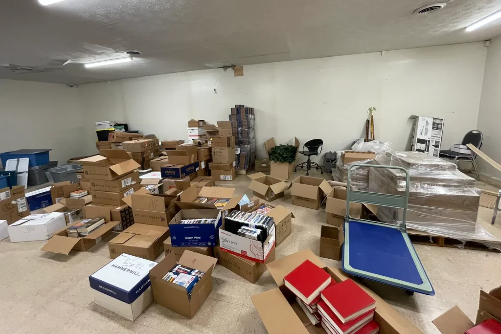 Boxed books and library materials stacked on pallets and carts in a temporary storage room.